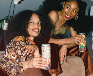 Two groovy women enjoy Oji Kola Original Kola and Pineapple Hibiscus flavored, Sparkling Energy Drinks at at a party